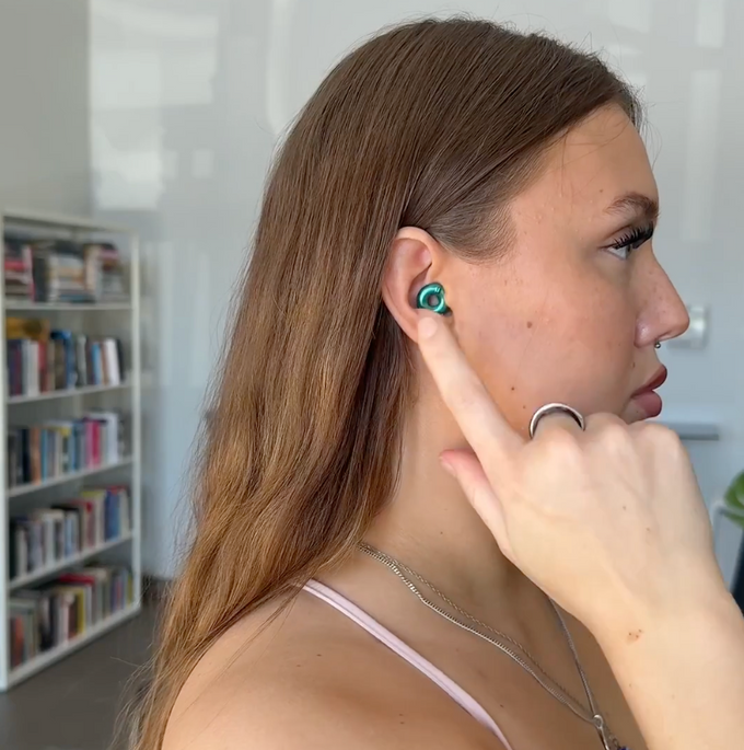 A woman with long light brown hair is shown in profile wearing Loop Switch 2 Earplugs, with her finger pointing toward it. She is wearing a silver ring and a thin necklace, with a blurred bookshelf in the background.