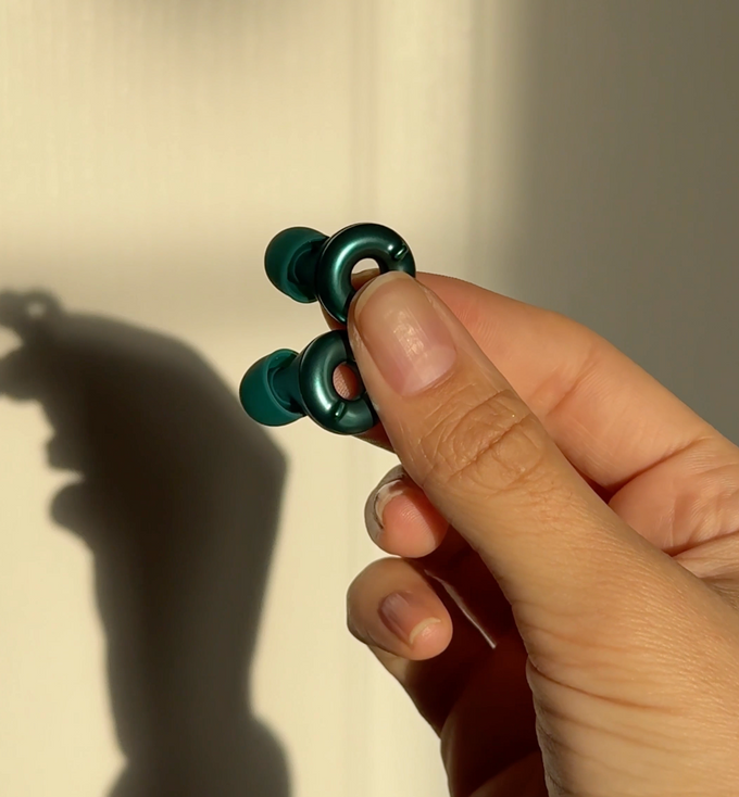 A close-up of a hand holding a pair of Loop Switch 2 Earplugs against a warm beige wall with a strong shadow.
