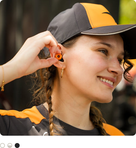 A close-up lifestyle shot of a woman wearing a black and orange baseball cap and a matching black and orange shirt, wearing McLaren x Loop Switch 2 earplugs.