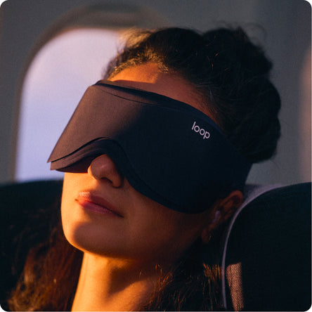 A woman wearing a black Loop sleep mask on an airplane, with warm sunlight highlighting her face and the mask.