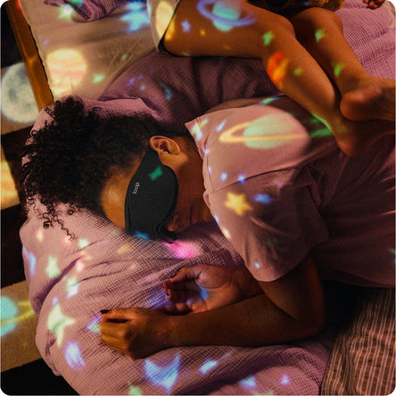 A person lying on a bed wearing a black sleep mask, with colorful planet and star-shaped light projections cast across the bedding and skin. 