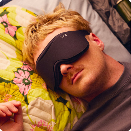 A man lying down wearing a black Loop sleep mask, with a yellow and pink floral quilt visible in the background.